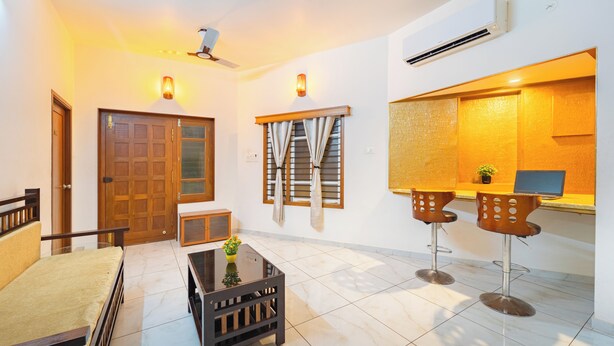 A living room with a sofa, a coffee table, and two bar stools. There is a wooden door, a window with curtains, and an air conditioning unit on the wall. - Roscotel Shara Inn Pondicherry (Puducherry)