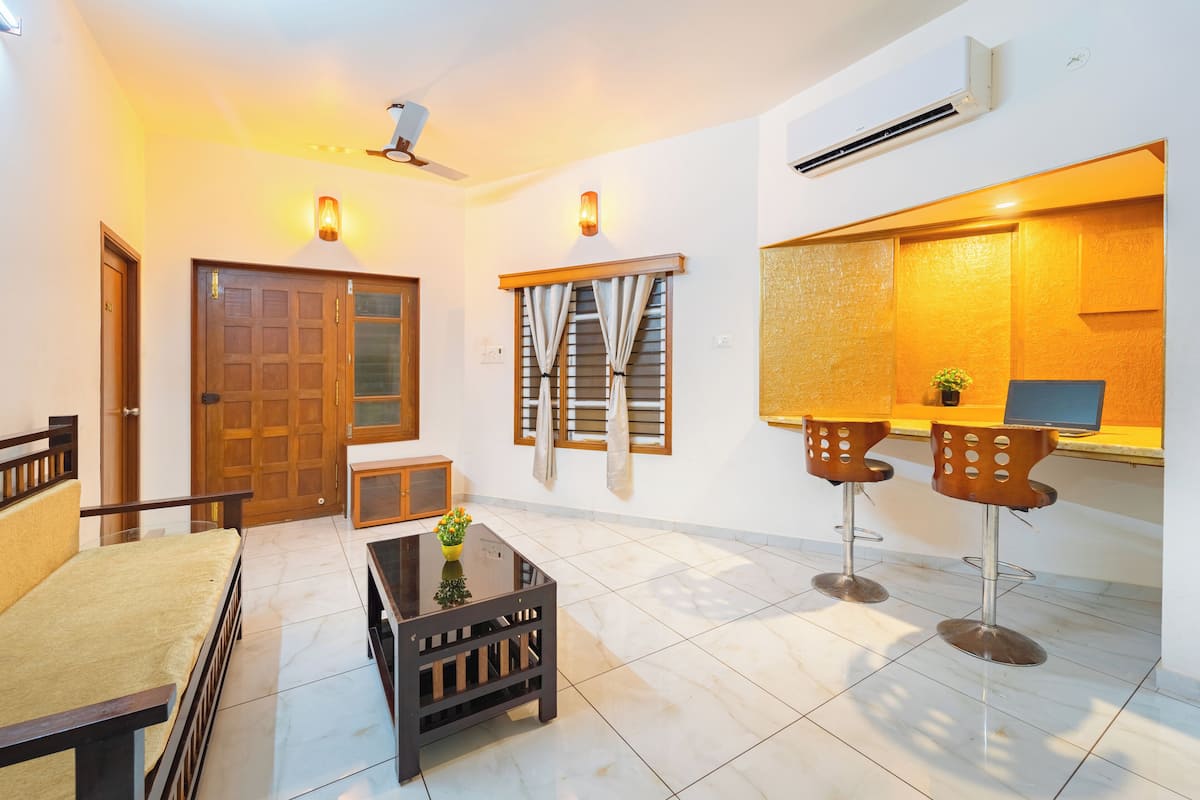 Un salon comprenant un canapé, une table basse et deux tabourets de bar. On y trouve une porte en bois, une fenêtre avec des rideaux et un appareil de climatisation fixé au mur.
