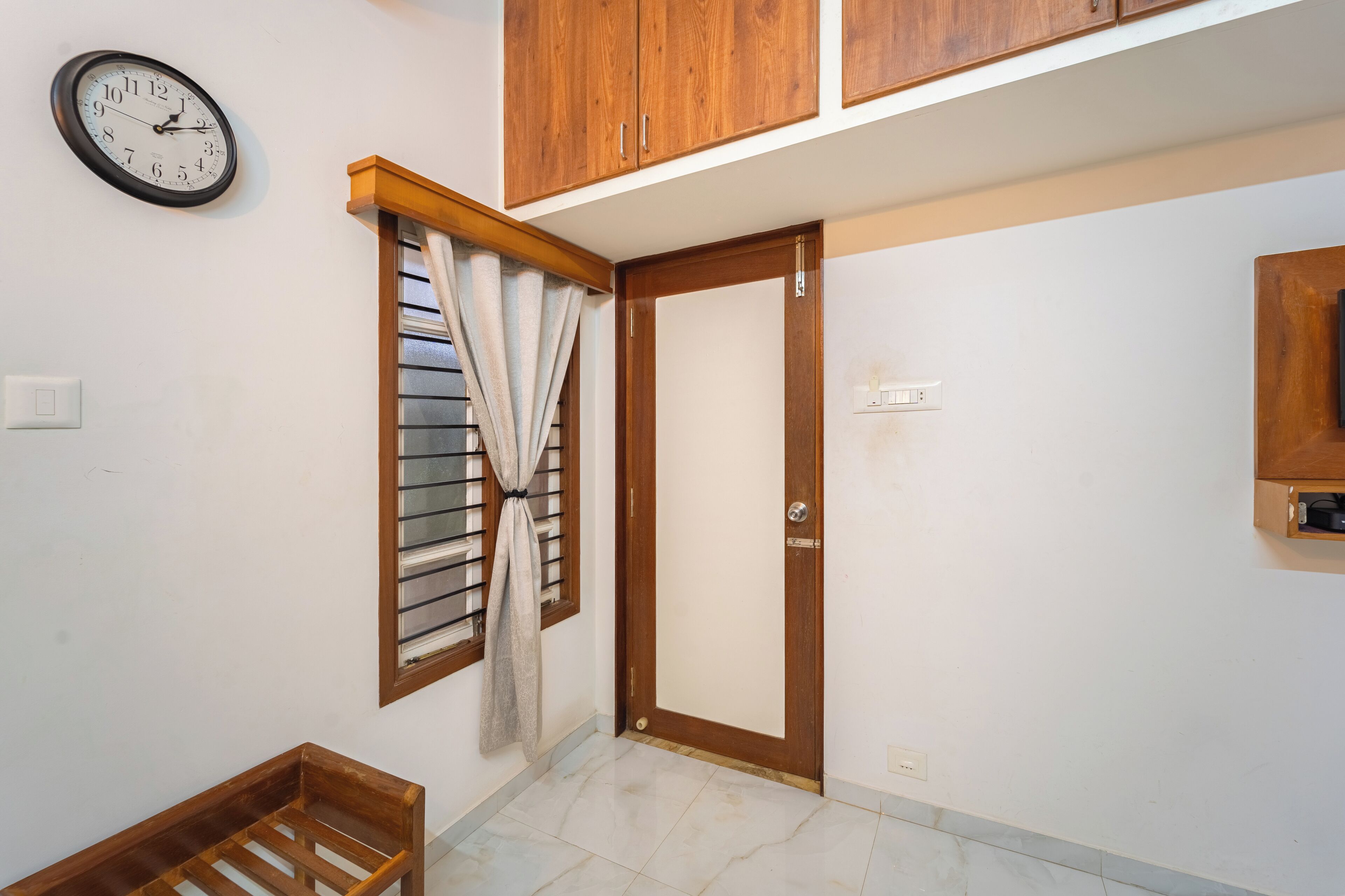 Una habitación con puerta de madera, una ventana con persianas, un banco de madera y un reloj sujeto a la pared.