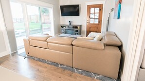 A modern living room with a large beige sectional sofa, a flat-screen TV mounted on the wall, and a wooden door. - Modern Cottage with Private Dock and large yard on Napa River (Napa)