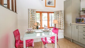 A dining area with a glass table, two pink chairs, a wooden ceiling, and a window with curtains. - The Forge (Taunton)