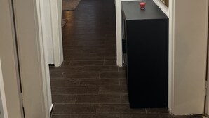 Een hal met een deur aan de linkerkant, een zwarte tafel met een rood voorwerp erop, en een raam met gordijnen op de achtergrond.