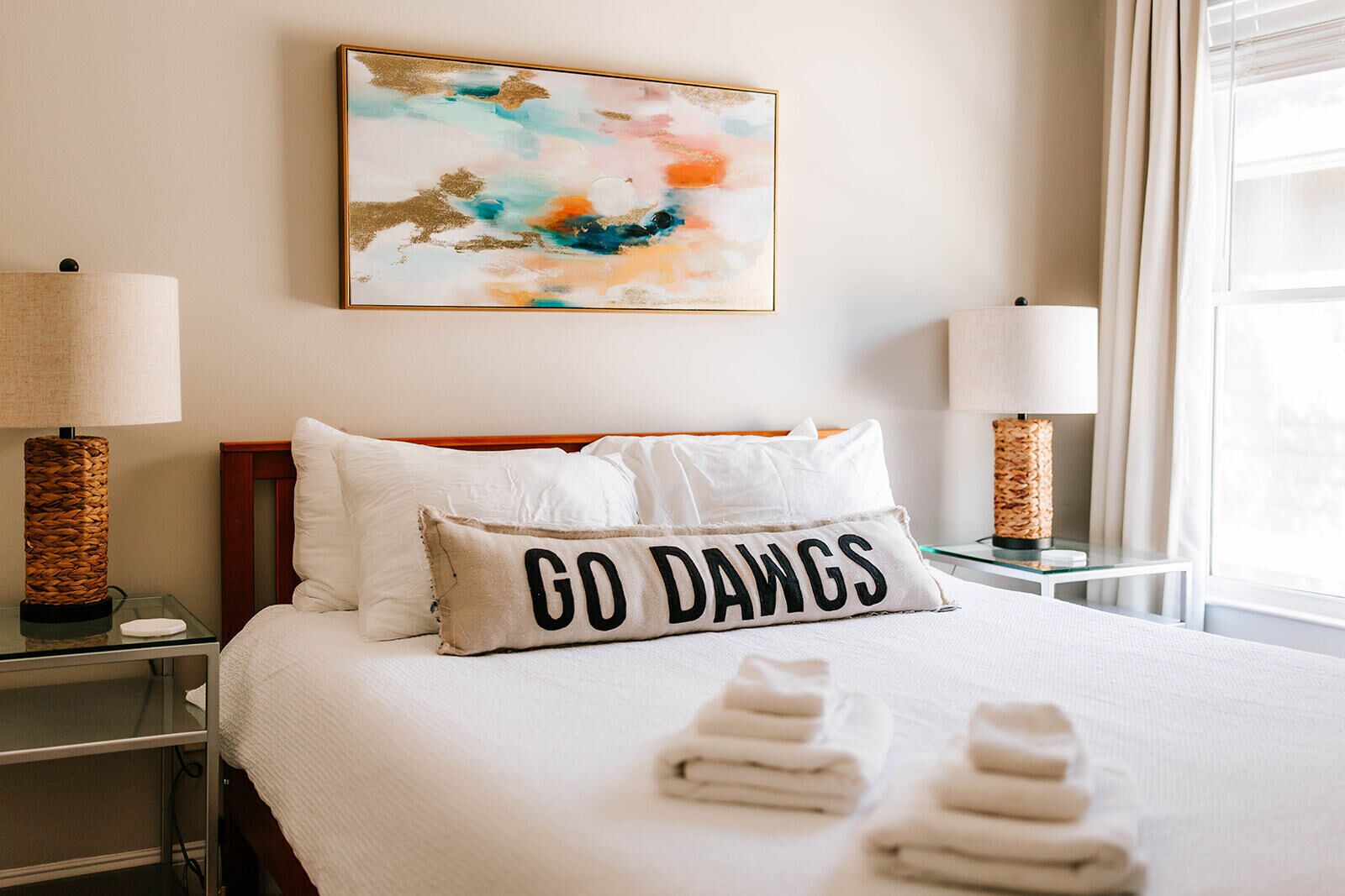 Hotelkamer met een bed, twee bedlampen, een wanddecoratie in abstractie stijl, en een kussen met de tekst 'GO DAWGS'.