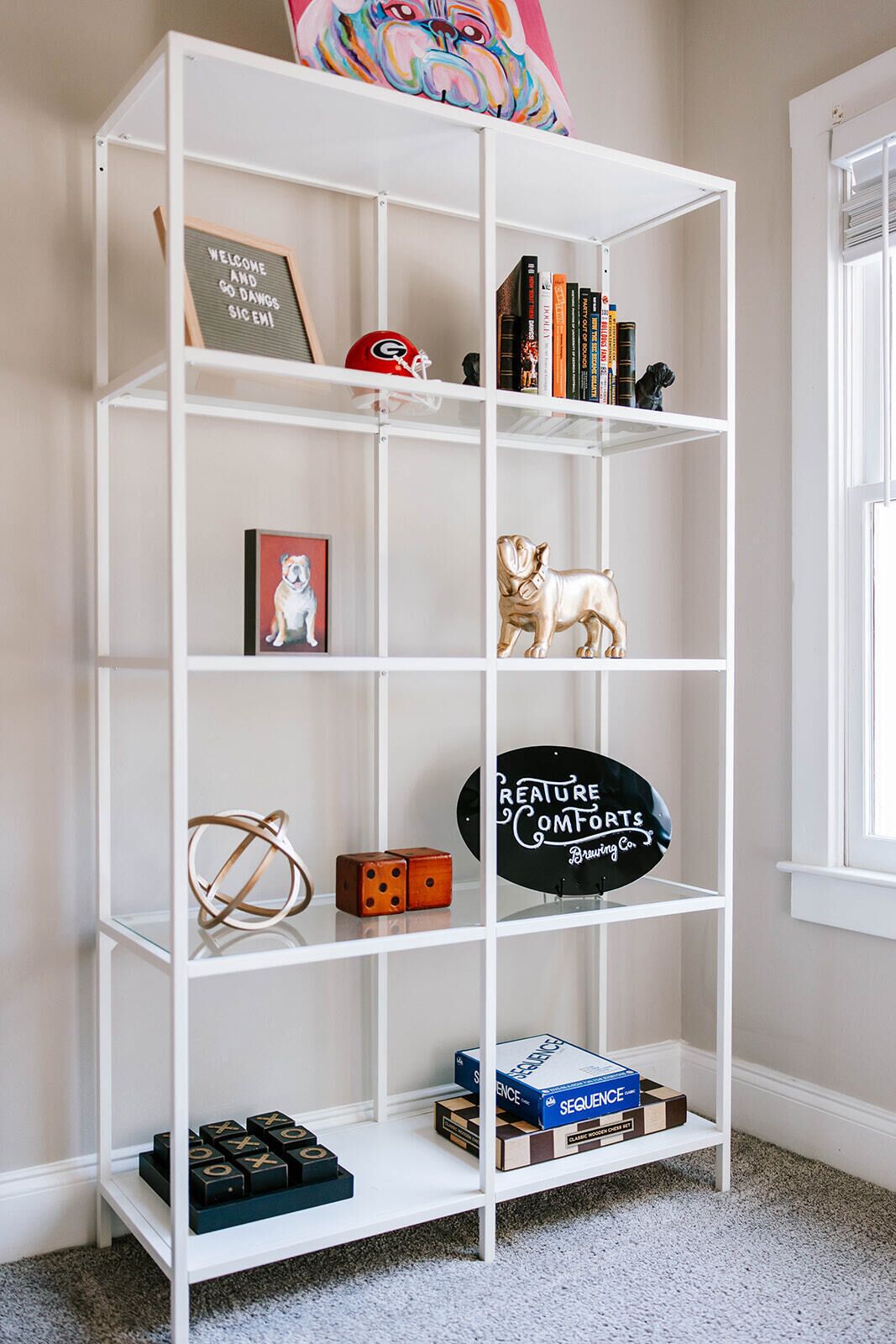 Een witte boekenkast met verschillende spullen, waaronder boeken, een bord met een welkomstboodschap, een voetbalhelm, een beeldje van een hond, dobbelstenen en een bord van een brouwerij.