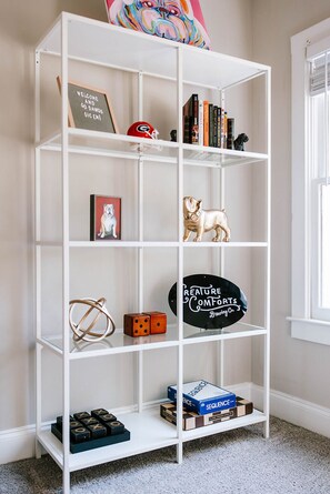 Une bibliothèque blanche contenant divers objets, notamment des livres, un panneau d’accueil encadré, un casque de football américain, une figurine de chien, des dés et un panneau publicitaire pour une brasserie.