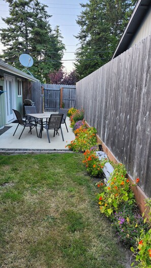 A backyard with a wooden fence, a patio table with chairs, and a variety of flowering plants.