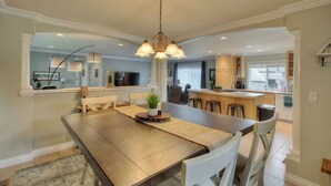 Un comedor amplio con una mesa de madera y sillas blancas, un lámpara colgante y vistas al área de la cocina.