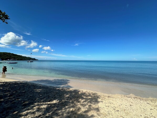 A beach with clear water, boats, and a person walking.