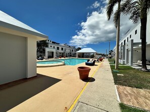 Zona de piscina con sillas de descanso y un área de estar techada, rodeada de edificios con un estilo arquitectónico moderno.