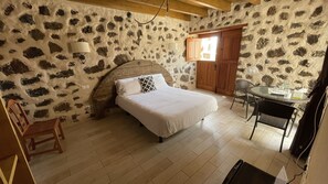 A bedroom with a stone-patterned wall, a wooden bed, a small table with chairs, and a window with a view. - Hotel Rural El Cabo (La Oliva)