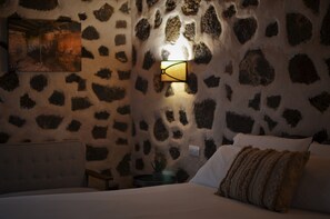 Una habitación con pared de piedra, cama con ropa de cama blanca y una lámpara fijada a la pared.