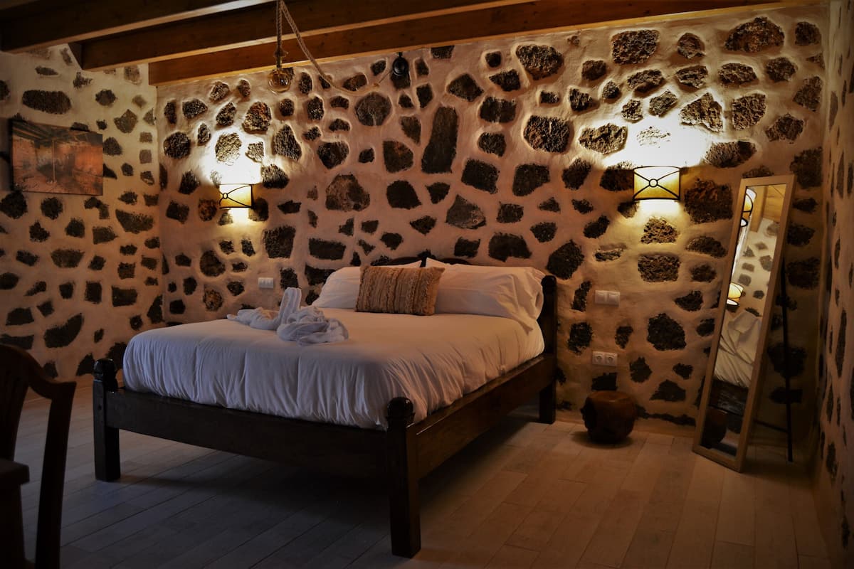 A bedroom with a stone wall, a wooden bed, two wall-mounted lights, and a mirror.