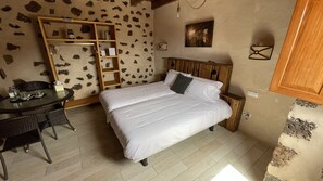 A bedroom with a wooden headboard, a bed with white bedding, a wooden wardrobe, a small table with chairs, and a wall with a stone-like pattern. - Hotel Rural El Cabo (La Oliva)