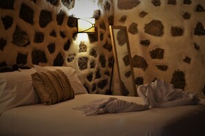 Un dormitorio con pared de piedra estampada, una cama con ropa blanca, una lámpara de pie de madera y una mesita de noche con lámpara.