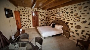 A bedroom with a stone wall, a wooden ceiling, a bed with white bedding, a wooden headboard, a desk with a chair, and a small table with bottles. - Hotel Rural El Cabo (La Oliva)