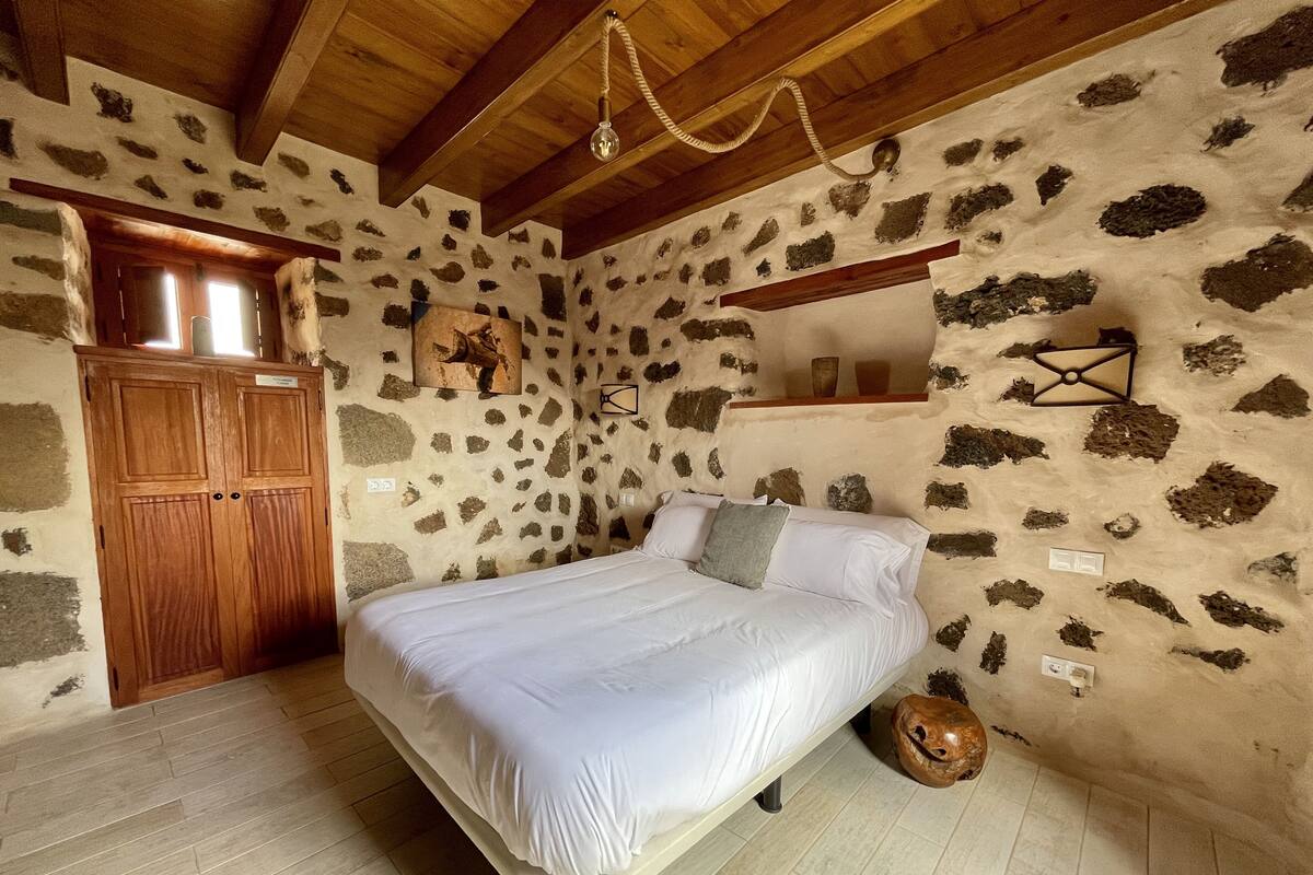 A bedroom with a stone wall, wooden ceiling beams, a bed, a wooden wardrobe, and a rustic decor piece.