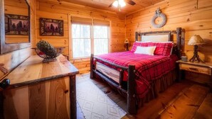 A cozy cabin bedroom with a wooden ceiling, a bed with red bedding, a ceiling fan, and a window with blinds. - Cozy lakeside cottage with outdoor haven for fishing, kayaking, and lounging (Ellijay)