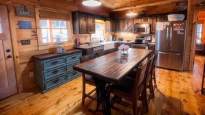 A wooden cabin interior with a central dining table, kitchen area with modern appliances, and a view of the outside. - Cozy lakeside cottage with outdoor haven for fishing, kayaking, and lounging (Ellijay)