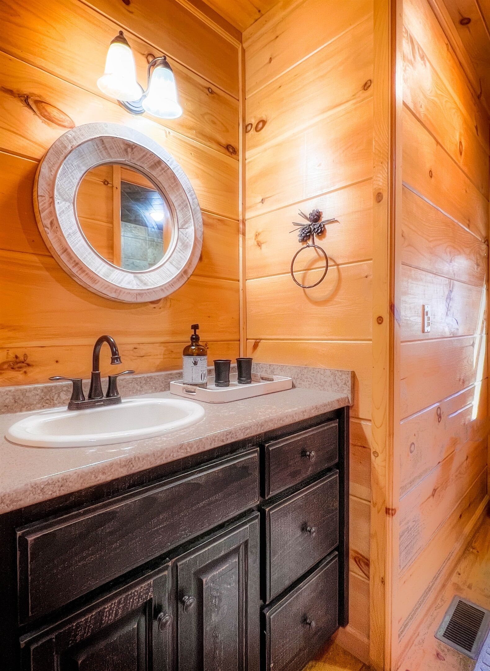 A wooden cabin bathroom with a round mirror, two sinks, a vanity with dark cabinets, and a pine wreath decoration.