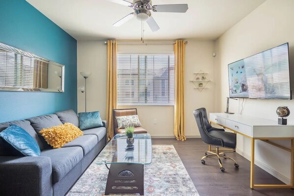 A modern living room with a grey sofa, a glass coffee table, a desk, and a large TV on the wall.