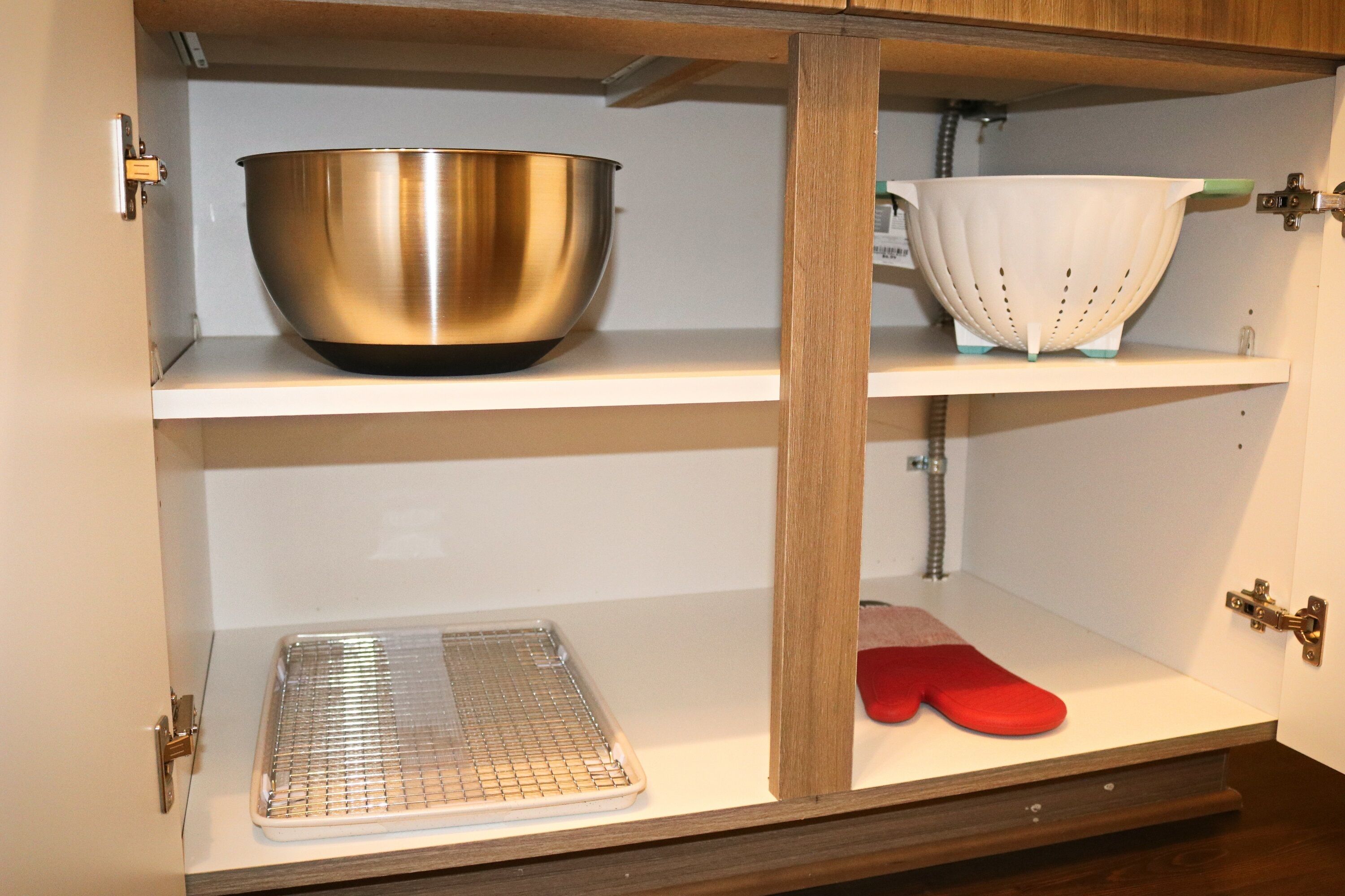 A kitchen cabinet with a stainless steel mixing bowl, a white colander, a baking tray, and red oven mitts.