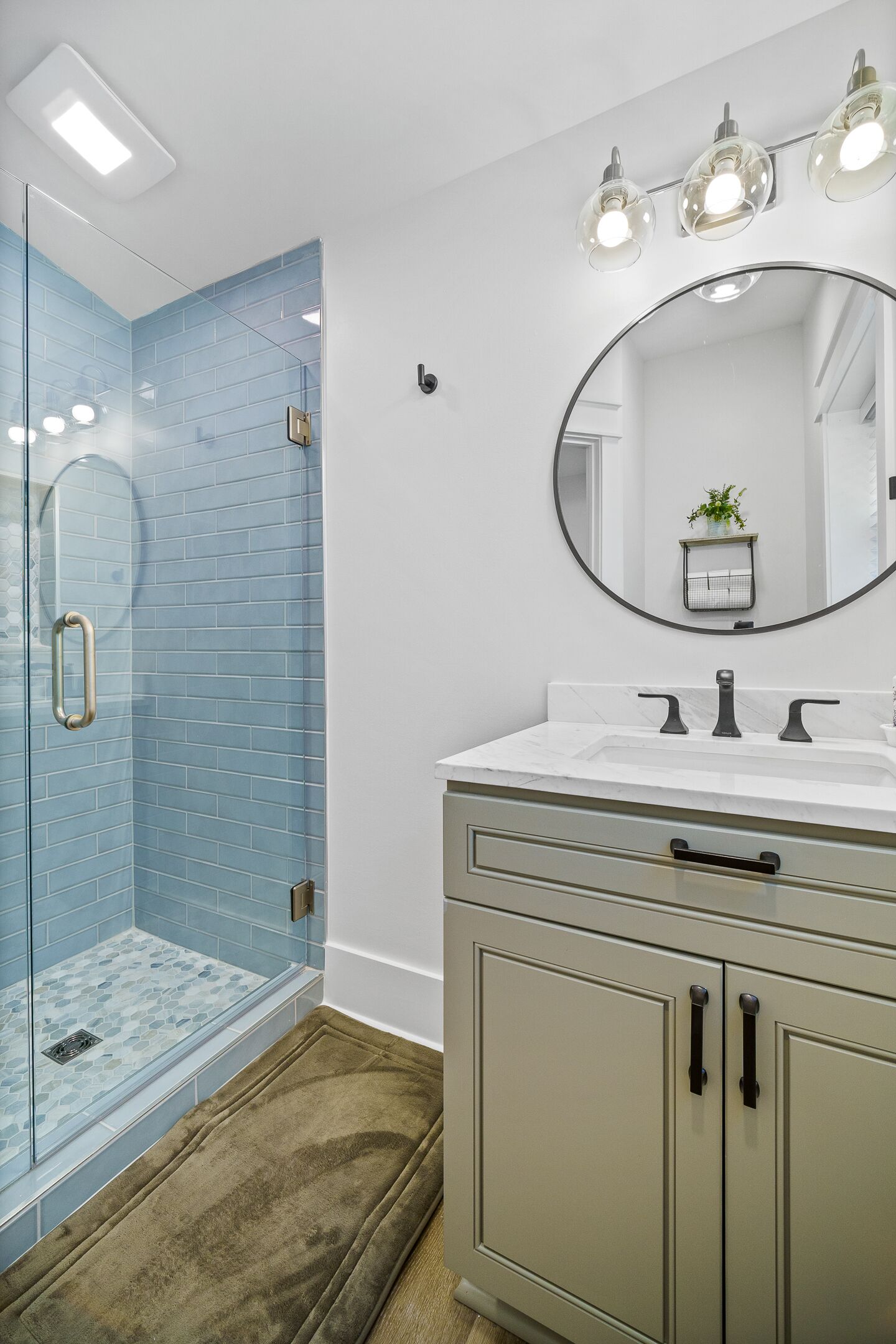Un baño con cabina de ducha de vidrio, espejo redondo y tocador con lavamanos y mueble.