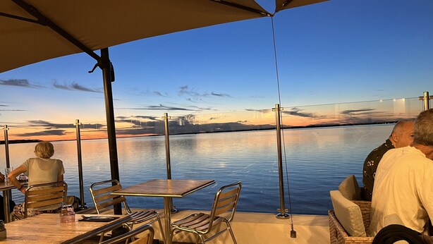 Un comedor con mesas y sillas, con vistas a una masa de agua tranquila y un horizonte urbano distante al atardecer.