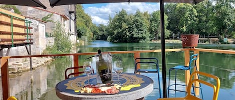 A table with a bottle of wine, a glass, and a plate of food on a boat deck with a view of a river and greenery.