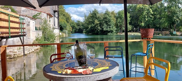 A table with a bottle of wine, a glass, and a plate of food on a boat deck with a view of a river and greenery.