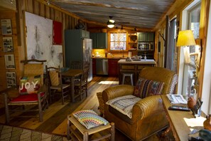Interior acogedor de una cabaña con un comedor de madera, un sofá con cojines de colores y una cocina con ventana y ventilador de techo.