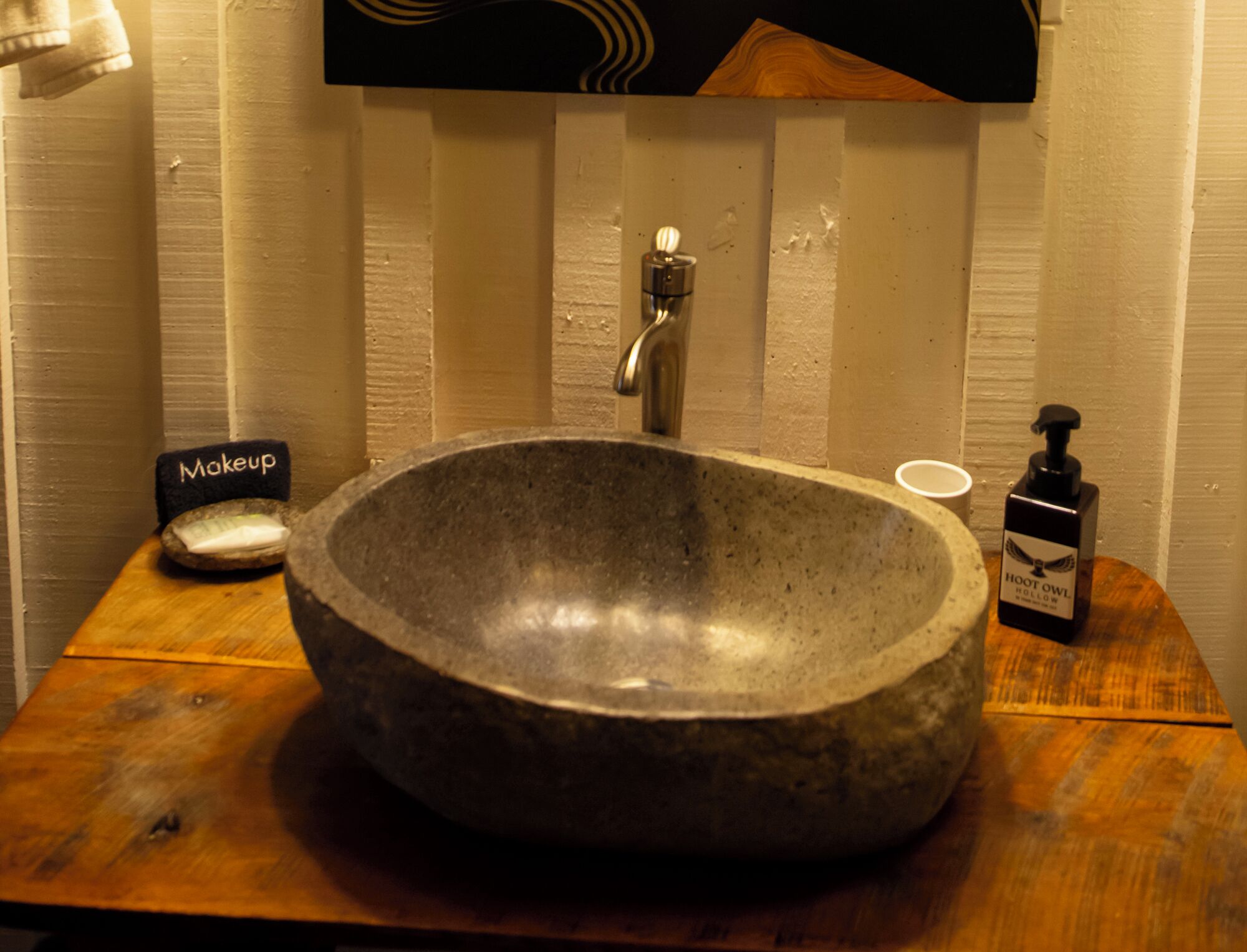 Un évier en pierre avec un robinet, un pinceau à maquillage et un distributeur de savon, le tout posé sur une table en bois dans une salle de bain.