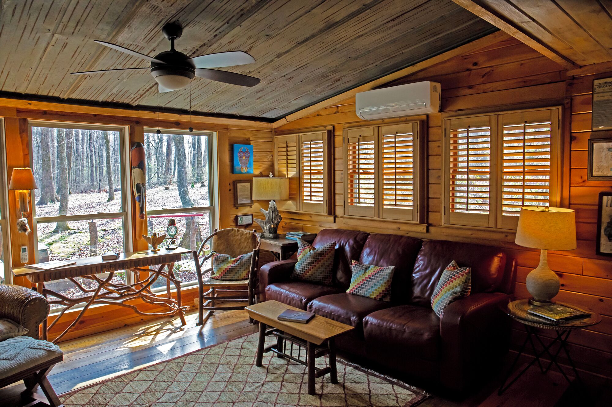 A cozy cabin interior with a wooden ceiling, a leather sofa, a dining table, and large windows with shutters.