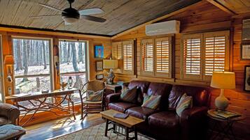 A cozy cabin interior with a wooden ceiling, a leather sofa, a dining table, and large windows with shutters.