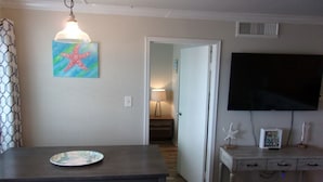 A dining area with a table and chairs, a painting of a starfish, a lamp, a TV, and a small cabinet with decorative items. - Sassy Seahorse - FRONT ROW - Beach Views 8 Guests (Galveston)