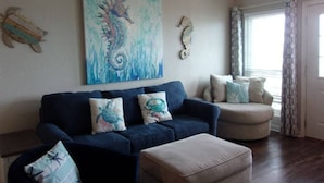 A living room with a dark blue sofa, beige armchair, and ottoman. A large seahorse painting adorns the wall. - Sassy Seahorse - FRONT ROW - Beach Views 8 Guests (Galveston)