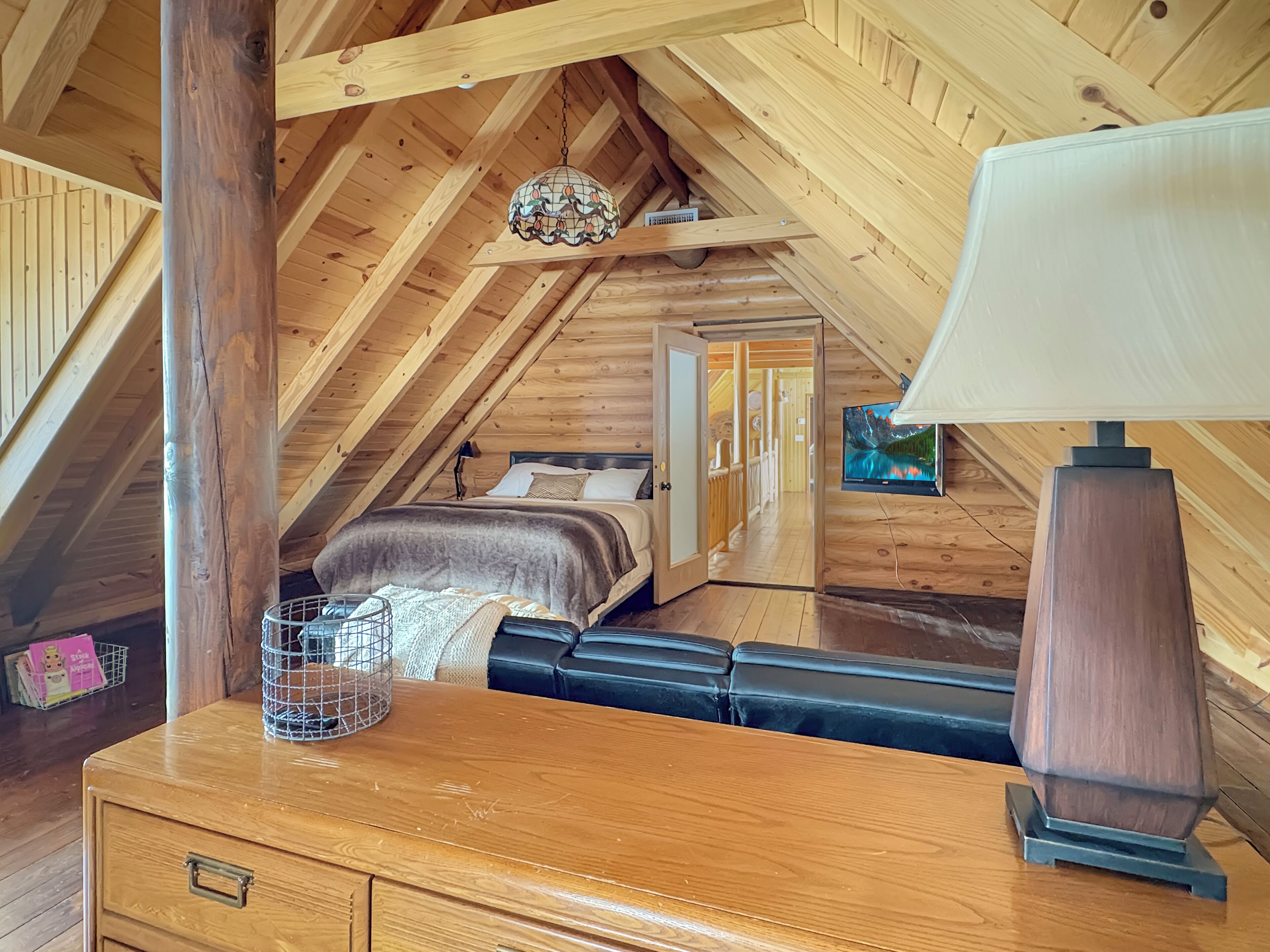 A cozy attic bedroom with a wooden ceiling, a bed with a grey blanket, a wooden dresser, and a TV on the wall.