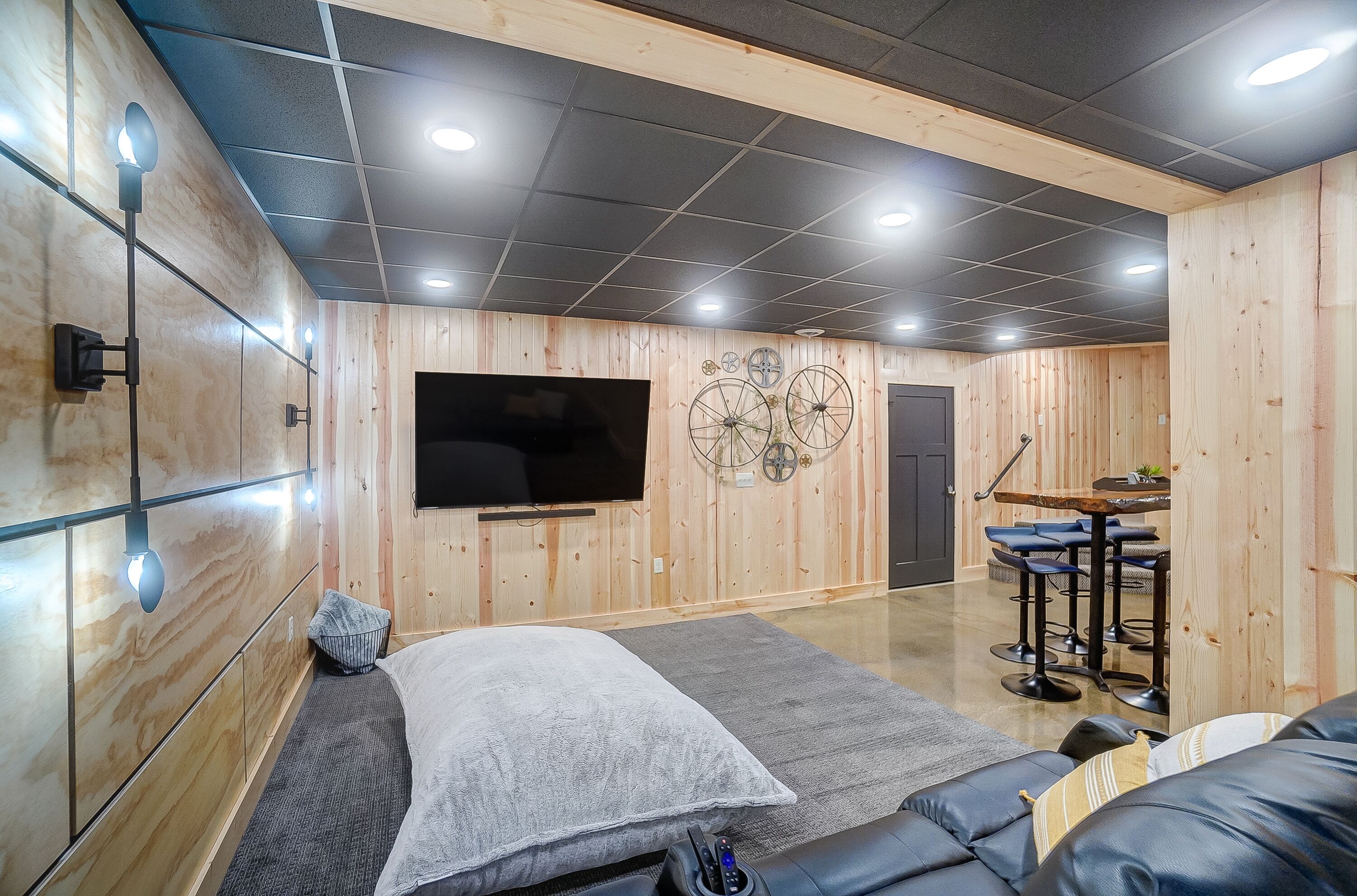 A modern room with a large TV, wooden walls, a sofa, and a small dining area with stools.