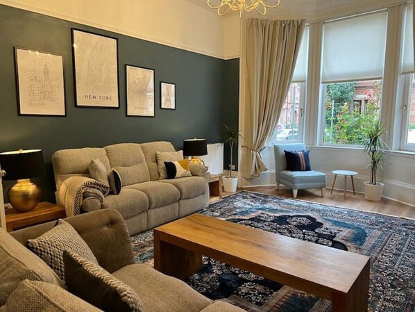 Un salon comprenant un canapé, des fauteuils, une table basse et un tapis. Les murs sont ornés de tableaux encadrés, il y a une lampe et une fenêtre avec des rideaux.