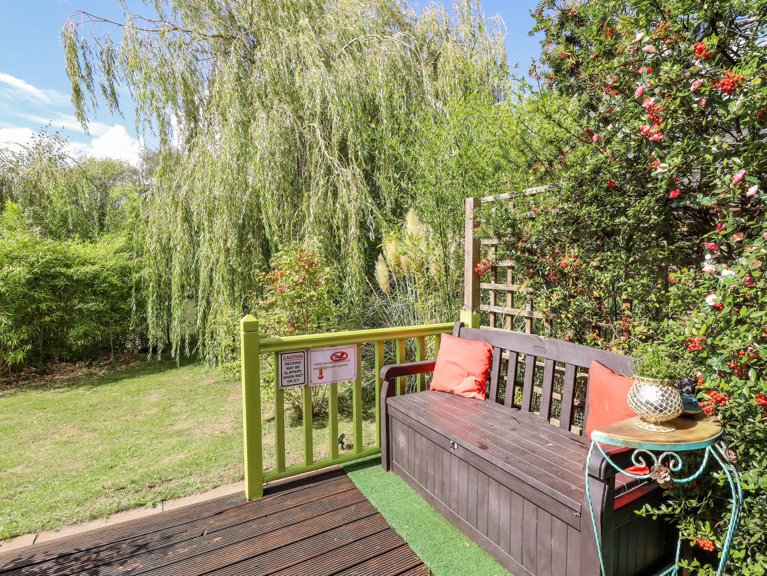 A wooden deck with a bench and a table, surrounded by lush greenery and a willow tree.