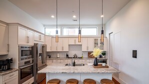 A modern kitchen with white cabinets, stainless steel appliances, a marble island with bar stools, pendant lighting, and a herringbone backsplash. - Luxury Midtown Houston Home w/ Rooftop Skyline Views (Houston)