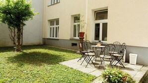 Um pátio externo com mesa, cadeiras, um vaso de planta e uma pequena árvore.