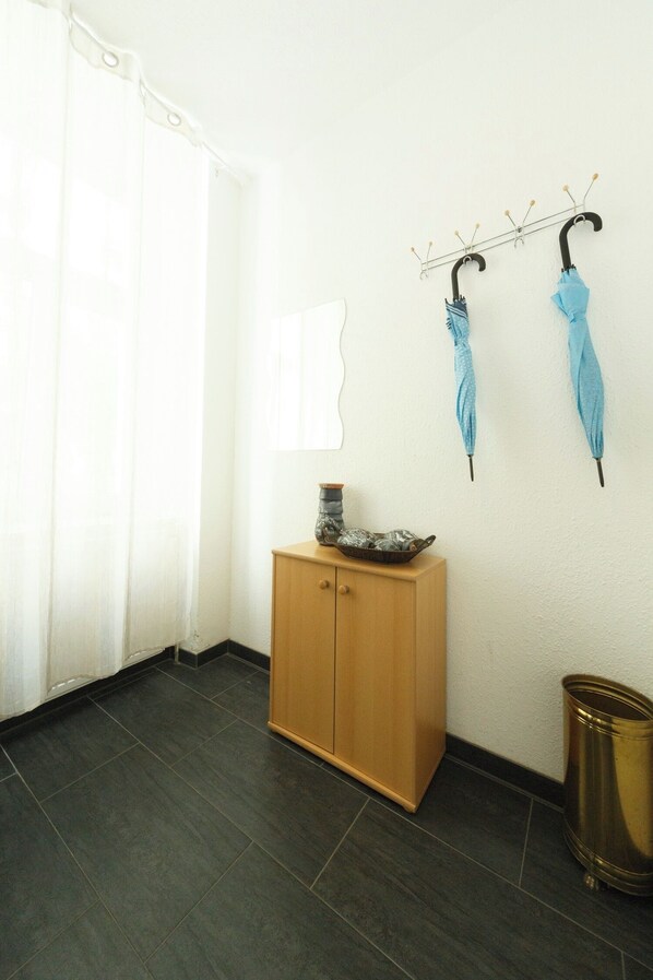 Een kamer met een houten kast, een metalen haak met twee paraplu's, een afvalbak en een wit gordijn.
