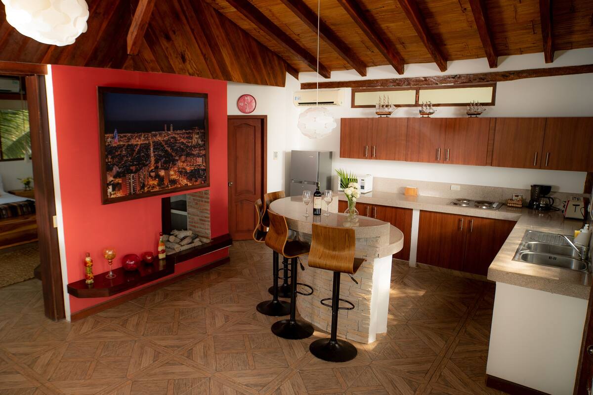 Une pièce spacieuse avec un plafond en bois, un mur d’accent rouge, un grand lit avec des coussins jaunes, un coin repas avec une table ronde et une cuisine avec des meubles en bois.