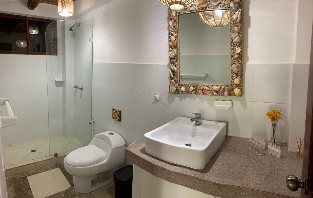 Ein Badezimmer mit Duschkabine aus Glas, Toilette, Waschbecken mit Spiegel und kleiner Blumenvase auf der Ablagefläche.