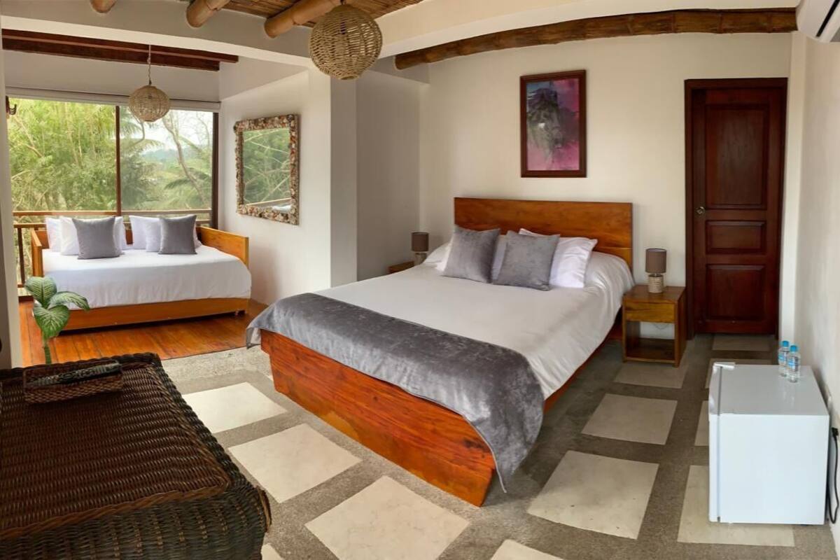 Une chambre avec un grand lit, un lit plus petit, une table de chevet, une commode, un réfrigérateur, une chaise en osier et une vue sur la verdure par la fenêtre.