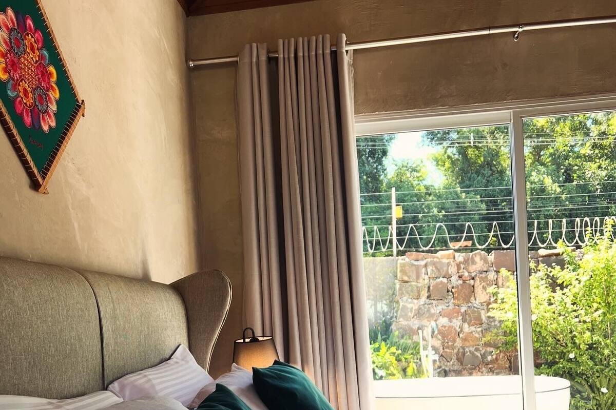 Een slaapkamer met een bed, kussens, een lamp en uitzicht op groen door een raam.