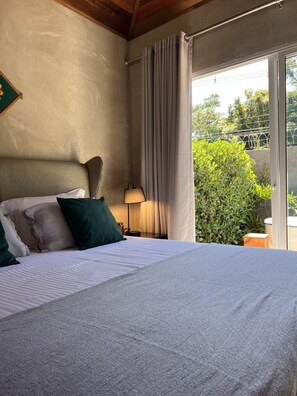 Une chambre à coucher avec un lit, des oreillers, une table de chevet, une lampe et une vue sur la verdure à l’extérieur.