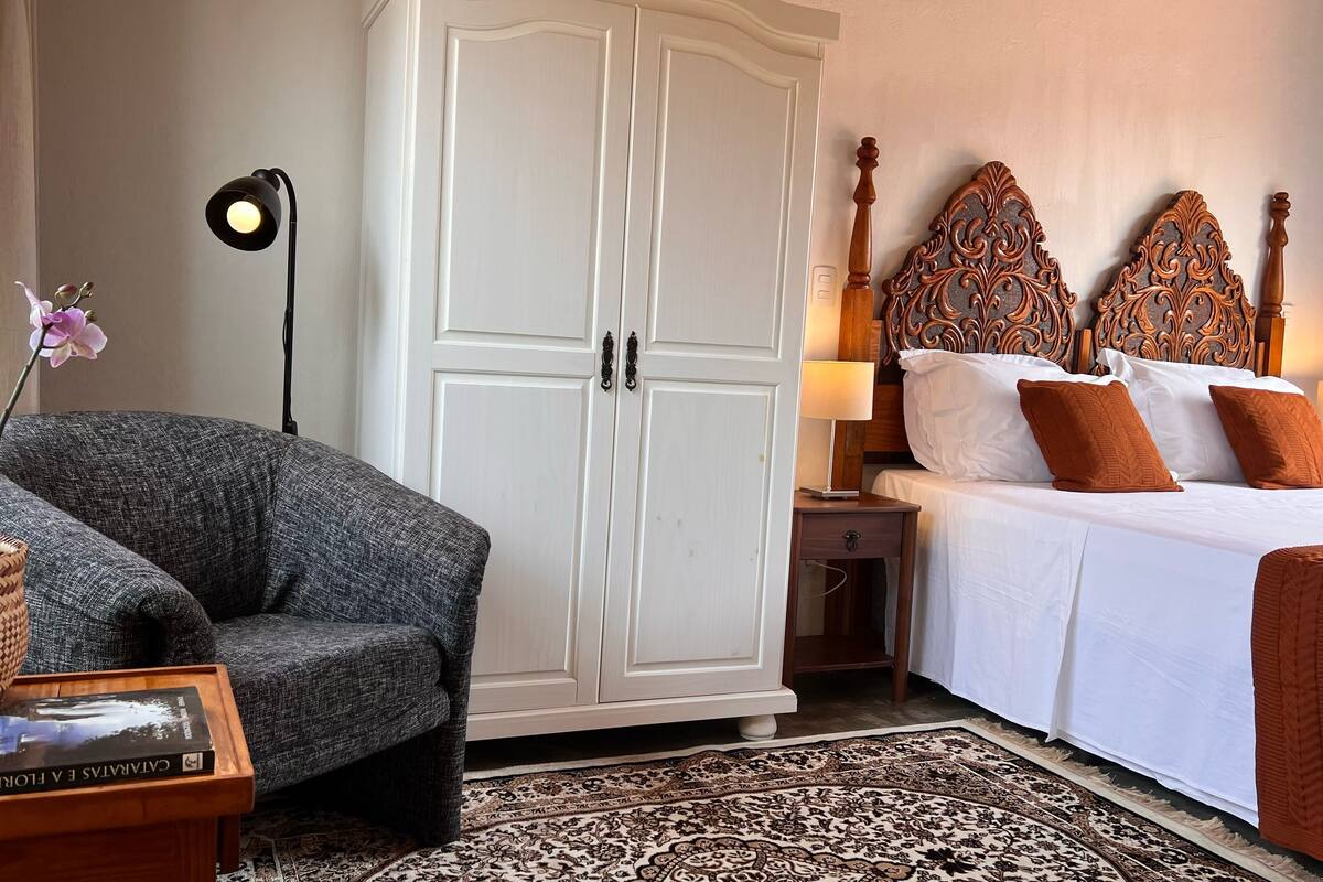 Een hotelkamer met een grote kledingkast, een bed met een versierd hoofdeinde, een grijze fauteuil, een nachtkastje met een lamp en een kamerbreed tapijt met dessin.
