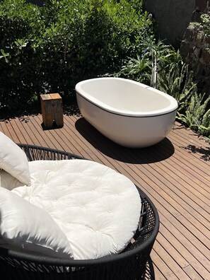 Un espace extérieur ensoleillé comprenant une grande baignoire blanche, un fauteuil en osier avec des coussins et un bloc de bois.