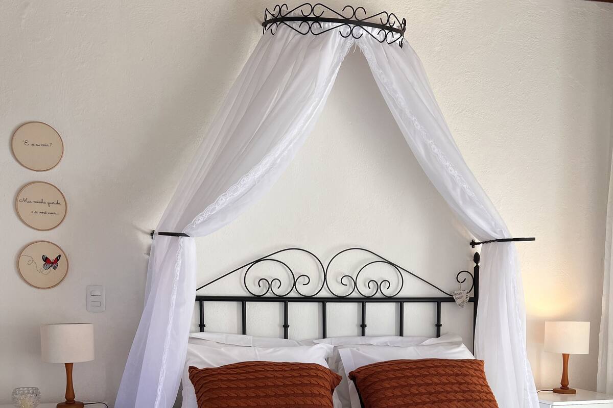 Een slaapkamer met een hemelbed, wit beddengoed en bruine kussens. Er staan twee nachtkastjes met lampen en een wand met decoratieve borden.